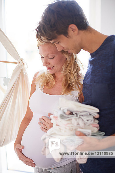 Schwangeres Paar hält Bauch und Stapel Babykleidung im Kinderzimmer