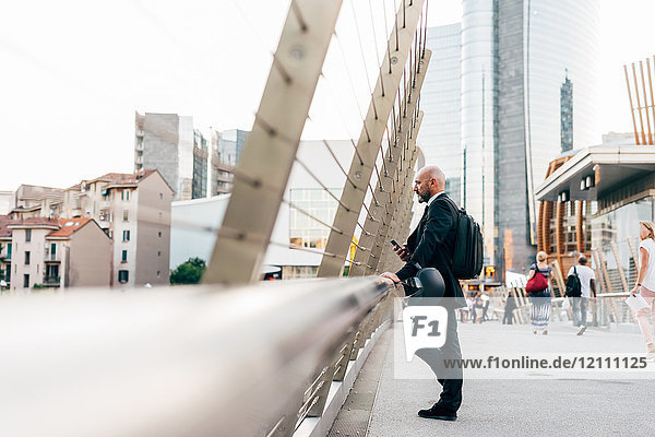 Reifer Geschäftsmann steht auf der Brücke  schaut auf die Aussicht  hält Smartphone