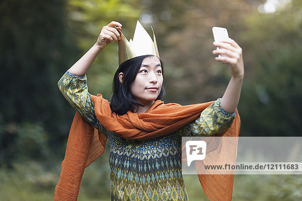 Junge Frau mit Krone  die Selfie durchs Handy nimmt  während sie im Park steht.