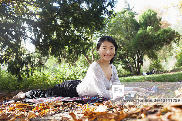Porträt einer jungen Frau  die auf einer Picknickdecke gegen Bäume im Park liegt und Yoga macht.