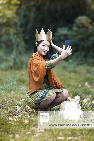 Junge Frau mit Krone  die Selfie durchs Handy nimmt  während sie auf dem Feld im Park hockt.