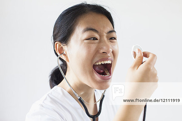 Frustrierter Arzt schreit mit offenem Mund in das Stethoskop gegen die weiße Wand.
