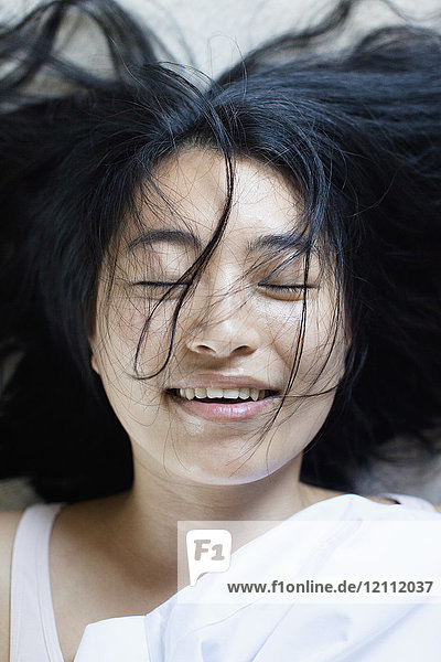 Hochwinkelansicht einer lächelnden jungen Frau mit geschlossenen Augen und unordentlichem Haar auf dem Bett liegend.