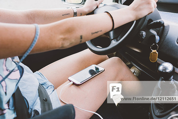 Mittelteil einer jungen Frau  die ein Fahrzeug mit einem Smartphone auf dem Schoß fährt