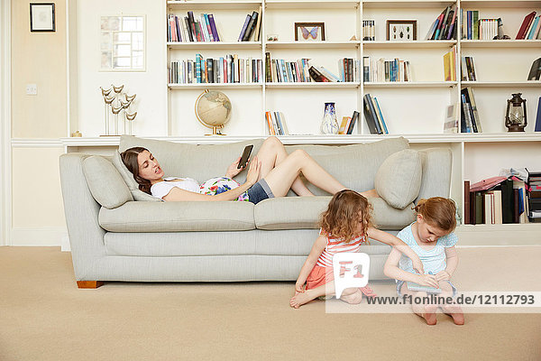 Schwangere Frau auf Sofa liegend mit Smartphone und Töchter auf Bodenzeichnung sitzend