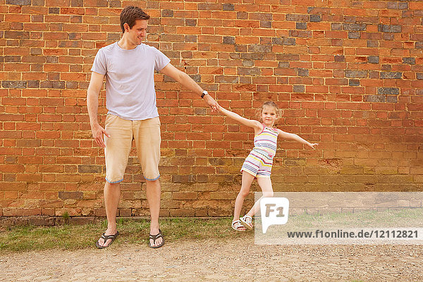Girl leaning sideways holding father's hand by brick wall