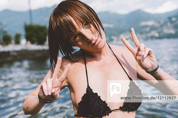 Portrait of young woman in bikini top making peace sign by Lake Como  Lombardy  Italy