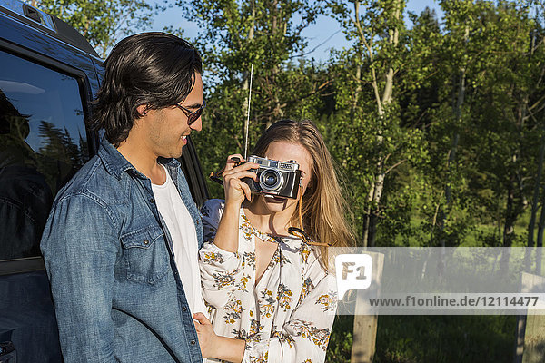 Junges Paar mit Kamera  das vor seinem Fahrzeug steht  während die junge Frau in der Ferne ein Foto macht; Edmonton  Alberta  Kanada