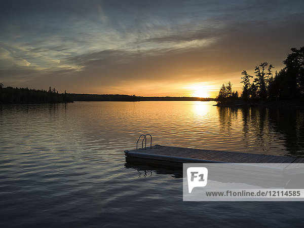 Sonnenuntergang über einem ruhigen See und silhouettierte Bäume entlang der Uferlinie mit einem Steg und einer Leiter im Vordergrund; Lake of the Woods  Ontario  Kanada