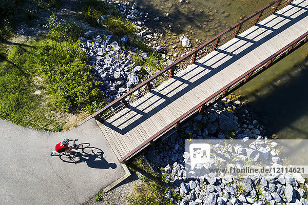View from directly above a cyclist in a red shirt going down a path and onto a bridge over a creek; Calgary  Alberta  Canada