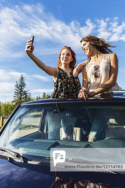 Zwei junge Frauen auf einem Roadtrip stehen auf dem Sonnendach eines Fahrzeugs und machen ein Selbstporträt mit einem Smartphone; Edmonton  Alberta  Kanada