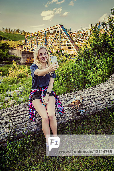 A young woman sits on a log in a park taking a self-portrait with her cell phone with a bridge and river in the background; Edmonton  Alberta  Canada