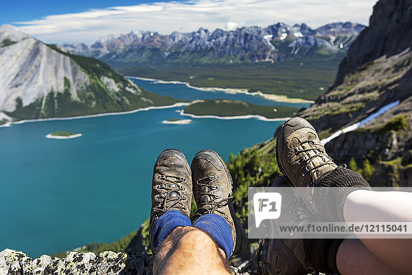Close-up of a couple's hiking boots over a cliff overlooking a colourful alpine lake and mountain range in the distance; Kananaskis Country  Alberta  Canada