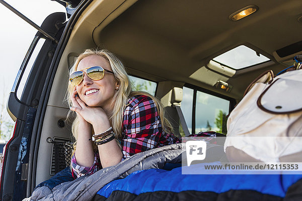 Eine junge Frau auf einer Autoreise liegt auf dem Rücksitz eines Fahrzeugs mit einem Schlafsack und schaut aus der offenen Tür; Edmonton  Alberta  Kanada