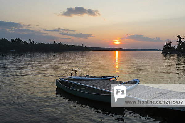 Sonnenuntergang über einem ruhigen See mit einem Steg  Kanu und Paddelbrettern im Vordergrund; Lake of the Woods  Ontario  Kanada
