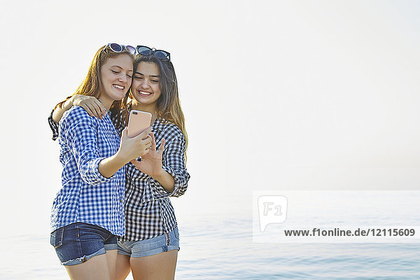 Two teenage girls take a self-portrait as they stand in an embrace at sunset at the lake  Woodbine Beach; Toronto  Ontario  Canada