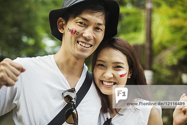 Portrait of man wearing hat and young woman with brown hair  Japanese flag painted on her cheek  smiling at camera.
