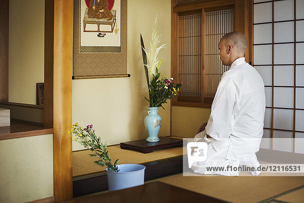 Side view of Buddhist monk with shaved head wearing white robe kneeling in front of vase with flowers.