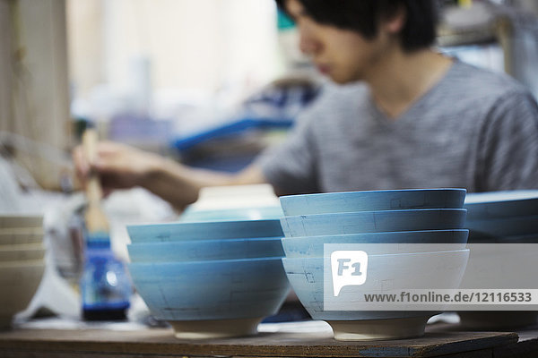 Nahaufnahme eines Mannes  der in einer japanischen Porzellanwerkstatt arbeitet und weiße Schalen mit blauer Glasur bemalt.