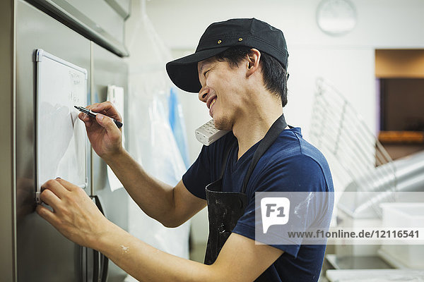 Man working in a bakery  wearing baseball cap and apron  writing note on small whiteboard  using phone and smiling.