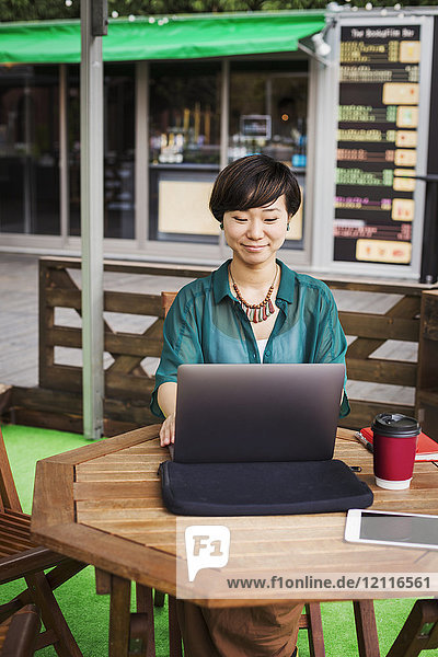 Frau mit schwarzen Haaren und grünem Hemd  die in einem Straßencafé vor einem Laptop am Tisch sitzt und arbeitet.