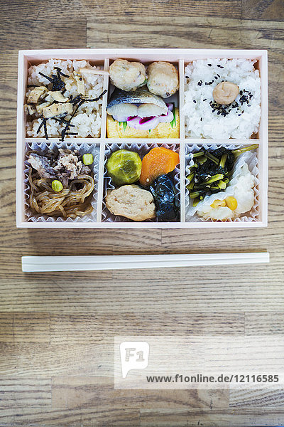 Hochwinkel-Nahaufnahme einer Bento-Kiste mit traditionellen japanischen Speisen und Essstäbchen auf einem Holztisch.