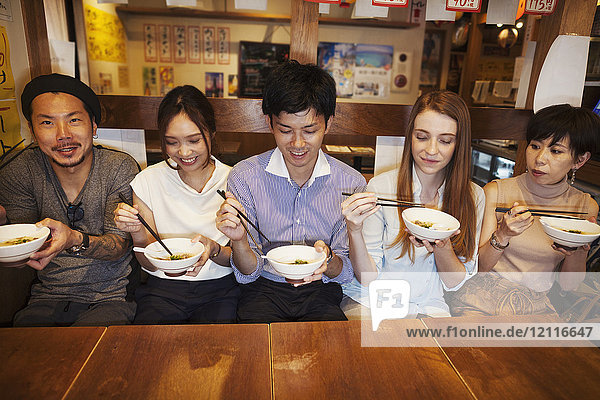 Five people sitting sidy by side at a table in a restaurant  eating from bowls using chopsticks.