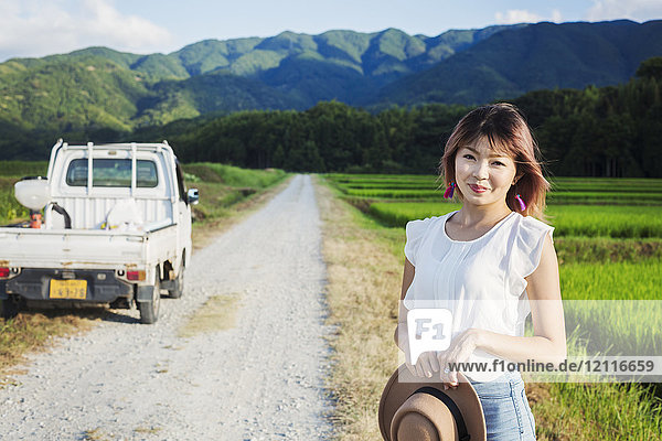 Eine junge Frau mit einem Sonnenhut in der Hand steht auf einer Straße an offenen Reisfeldern.