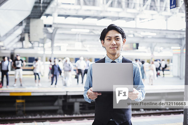 Geschäftsmann in blauem Hemd und Weste steht auf dem Bahnsteig des Bahnhofs  hält Laptop in der Hand und schaut in die Kamera.