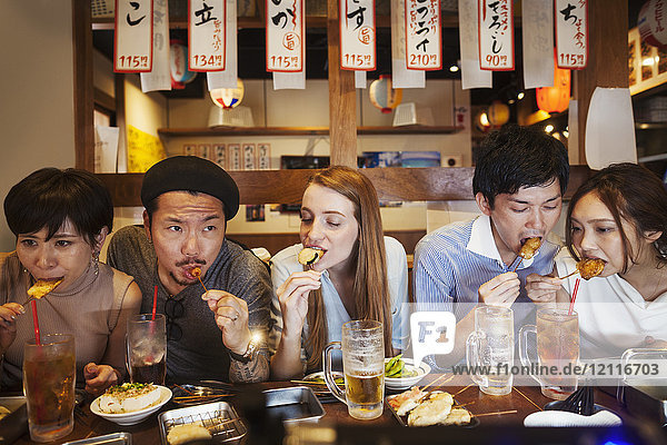Five people sitting sidy by side at a table in a restaurant  eating from skewers.