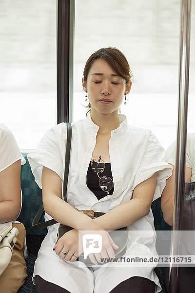 Frau mit schwarzen Haaren und weißem Hemd im Nahverkehrszug sitzend  Augen geschlossen.