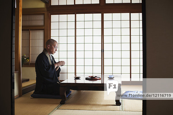Side view of Buddhist monk with shaved head wearing black robe kneeling indoors at a table  holding bowl of tea.