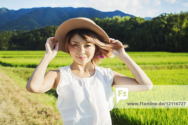 Eine junge Frau hält einen Strohhut auf dem Kopf  die Haare wehen im Wind  auf freiem Feld an Reisfeldern.