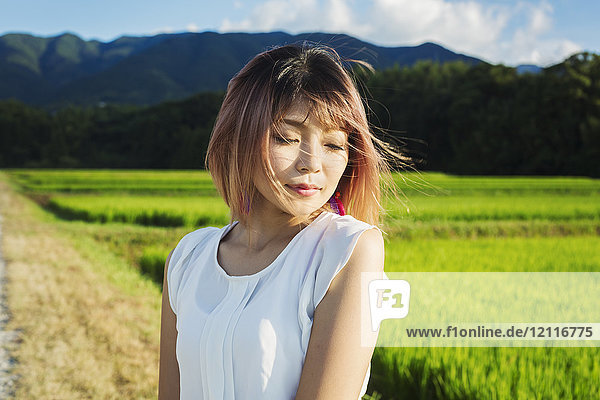 Eine junge Frau in einem weißen Hemd  mit vom Wind verwehten Haaren  steht im Freien an Reisfeldern.
