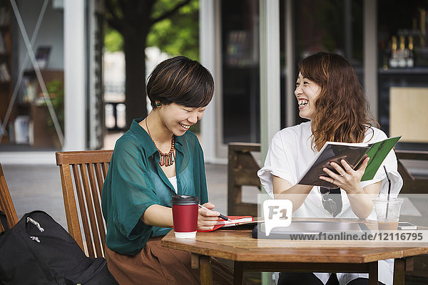 Two women with black hair wearing green and white shirt sitting at table in a street cafe  holding digital tablet  smiling.