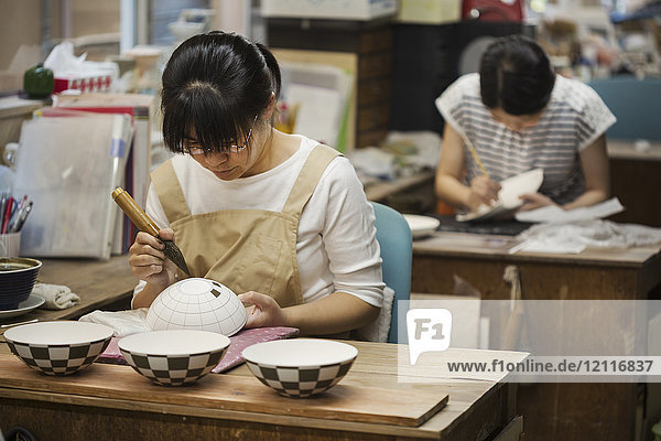 Frau  die in einer japanischen Porzellanwerkstatt arbeitet und mit dem Pinsel geometrische Muster auf weiße Schalen malt.