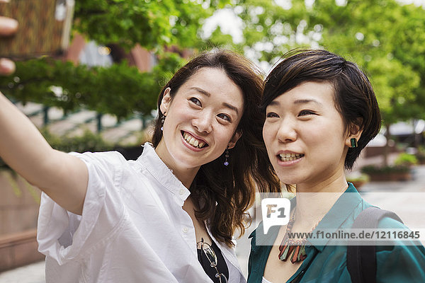 Two women with black hair wearing white and green shirt standing outdoors  taking selfie with mobile phone  smiling.