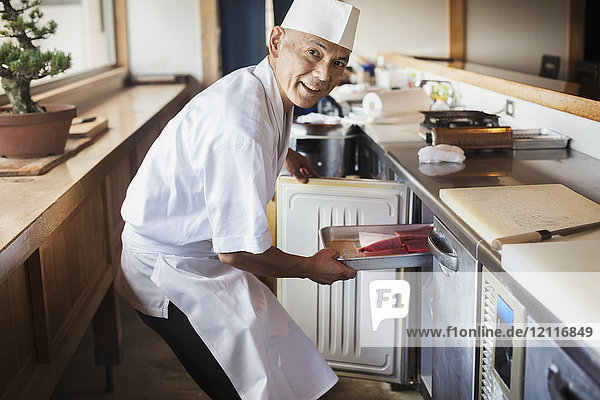 Chefkoch  der in einem japanischen Sushi-Restaurant an der Theke arbeitet und ein Metalltablett mit Fisch in den Kühlschrank stellt.