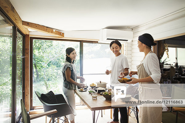 Mann und zwei lächelnde Frauen stehen um einen Tisch mit Tellern und Schüsseln mit Essen.
