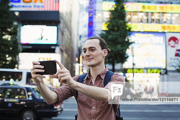 Ein junger Mann  ein kaukasischer Besucher Tokios  der auf der Straße mit seinem Smartphone unterwegs ist und Fotos macht.