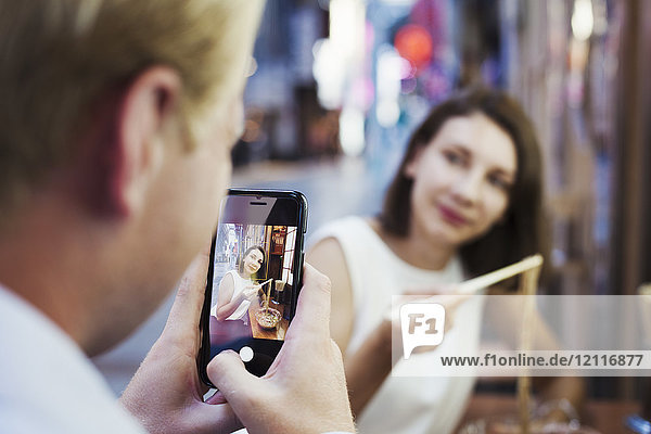 Man taking picture with smartphone of woman sitting at a table in an asian restaurant  using chopsticks  eating noodles.