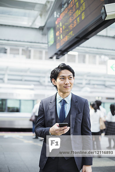 Geschäftsmann im Anzug  steht auf dem Bahnsteig und hält ein Mobiltelefon in der Hand.