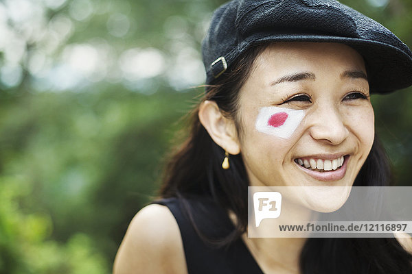 Porträt einer lächelnden jungen Frau mit schwarzen Haaren und flacher Mütze  auf der Wange die japanische Flagge gemalt.