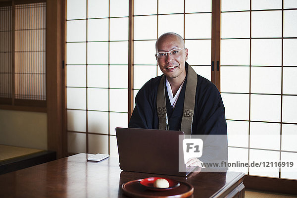Buddhist monk with shaved head wearing black robe sitting indoors at a table  using laptop computer  smiling at camera.