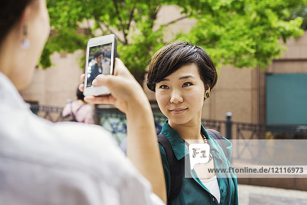 Two women with black hair wearing white and green shirt standing outdoors  taking picture with mobile phone  smiling.