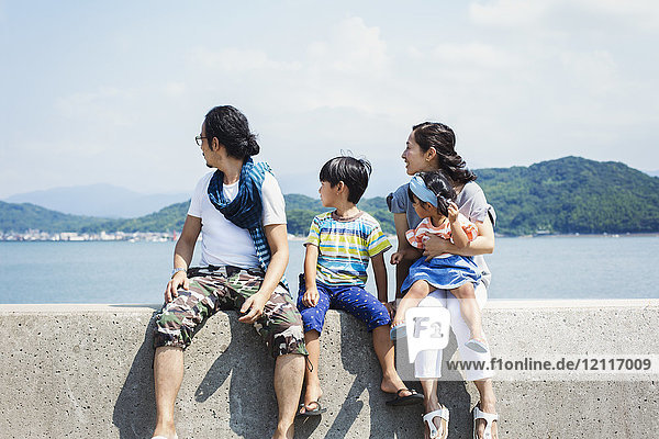 Eine Familie  ein Mann  ein Junge und eine Frau mit einem jungen Mädchen auf dem Schoß  die Seite an Seite auf einer Mauer am Meer sitzen.