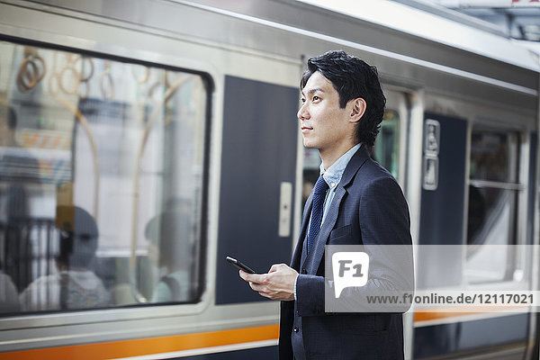 Geschäftsmann im Anzug steht am Bahnsteig und hält ein Mobiltelefon in der Hand.