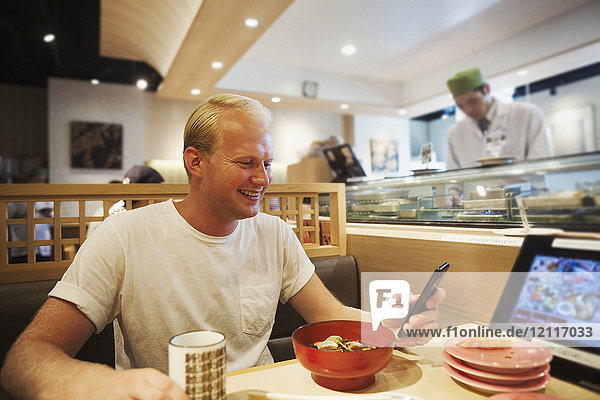 Junger Mann sitzt an einem Tisch in einem asiatischen Fast-Food-Restaurant  schaut auf sein Smartphone und lächelt.