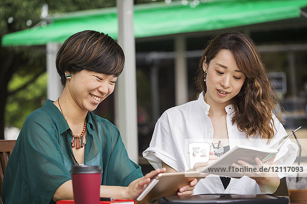 Zwei Frauen mit schwarzen Haaren in grün-weißem Hemd sitzen am Tisch in einem Straßencafé  halten ein digitales Tablett in der Hand und lächeln.