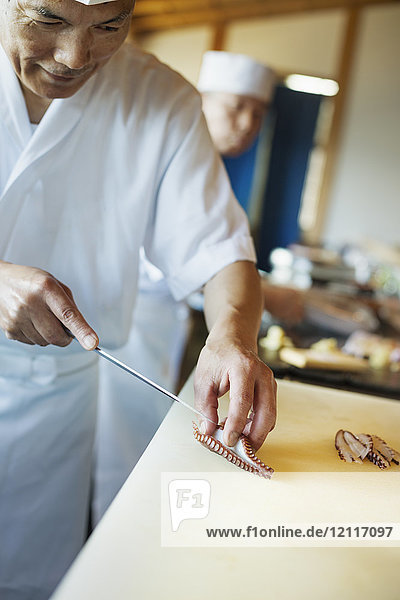 Zwei Köche arbeiten an einer Theke in einem japanischen Sushi-Restaurant und schneiden Tintenfisch-Tentakel in Scheiben.